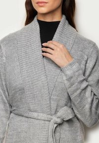 Cardigan gri tricotat cu guler tip șal, textură bobitată și o curea legată în talie; material moale cu un aspect confortabil.