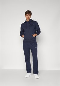 Marineblauer Baumwoll-Hoodie mit Kordelzug, Fronttasche und Logo. Kombiniert mit passenden Jogginghosen und weißen Sneakern. Glatte Textur.