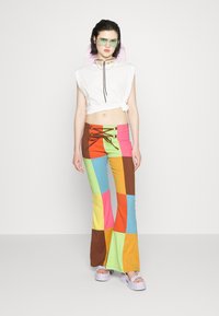 Top branco curto com detalhe knotted, calças largas multicoloridas com grandes retalhos geométricos e sapatos de plataforma grossa. Óculos de sol verdes.