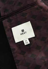 Burgunderfarbener Stoff mit geometrischem Muster, auf einem dunklen Hintergrund, mit einem sichtbaren weißen Etikett, auf dem "NEXT" und die Größe "M" aufgenäht sind.