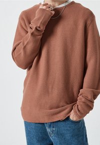 Person, der ein strukturiertes braunes Langarmhemd, blaue Jeans, eine silberne Kette und einen Ring trägt, mit einer Hand in der Tasche und der anderen nahe am Gesicht.