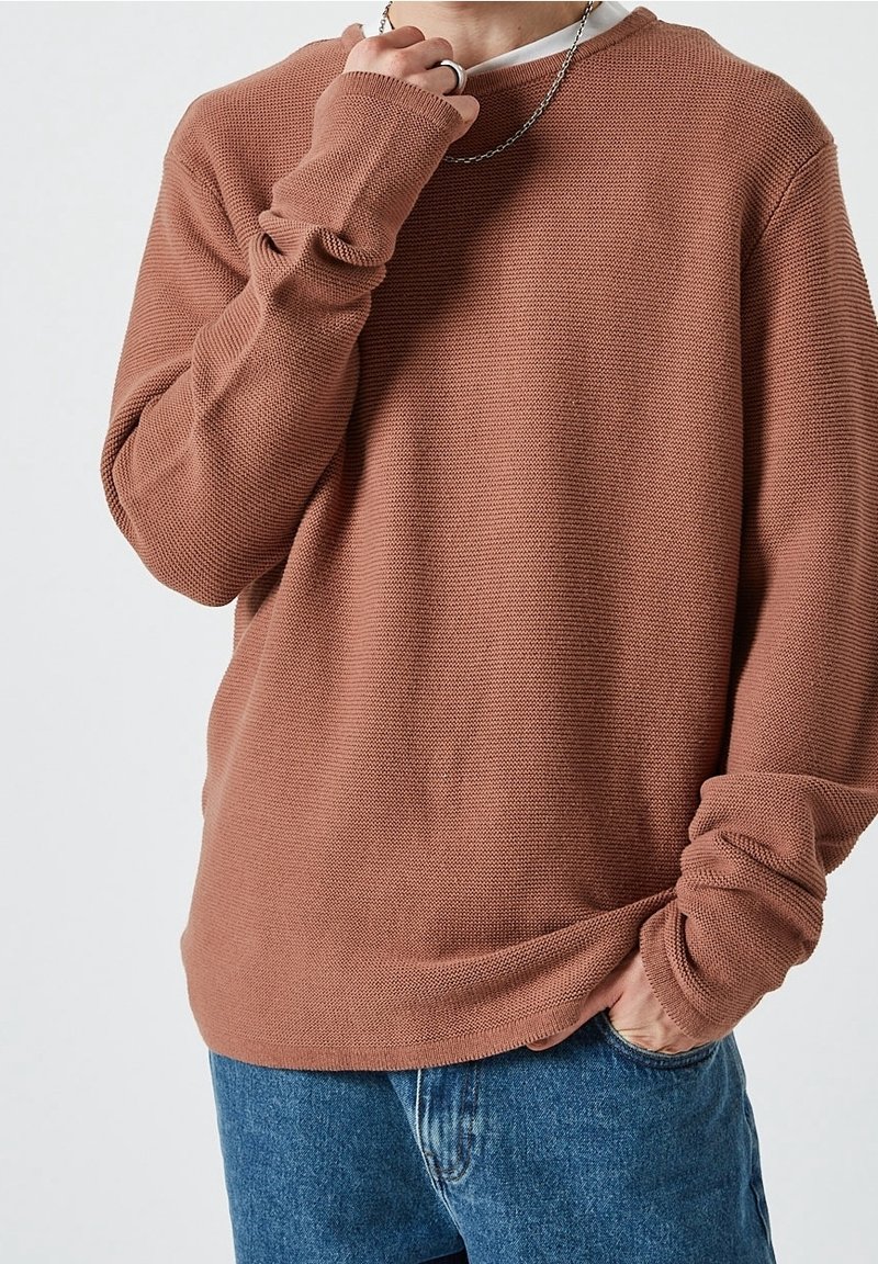 Person, der ein strukturiertes braunes Langarmhemd, blaue Jeans, eine silberne Kette und einen Ring trägt, mit einer Hand in der Tasche und der anderen nahe am Gesicht.