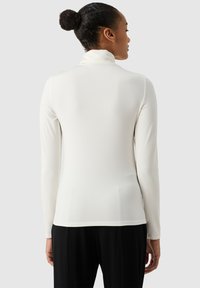 Top bianco a maniche lunghe con colletto alto, realizzato in un tessuto morbido. Presenta una silhouette aderente e un design minimalista. Pantaloni neri visibili sotto.