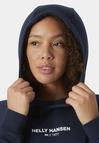 Hoodie navy realizzato in tessuto morbido con cappuccio regolabile. Presenta il nome del marchio "Helly Hansen" e il logo in bianco sul davanti.