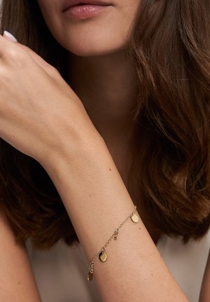 Nahaufnahme eines Handgelenks einer Frau, die ein filigranes Armband aus einer goldenen Kette mit kleinen flachen runden Anhängern trägt.