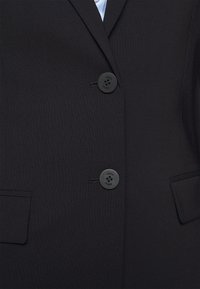 Svart blazer med två knappar, fickor med slag och ett mjukt, texturerat tyg. Knapparna har graverat varumärkesnamn, vilket ger detaljer till designen.