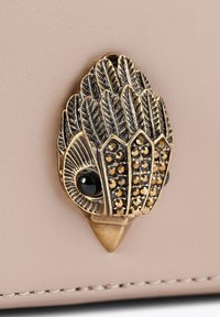 Pochette en cuir beige avec une fermeture décorative en bronze ressemblant à une plume, présentant des détails complexes et des accents de pierres précieuses noires.