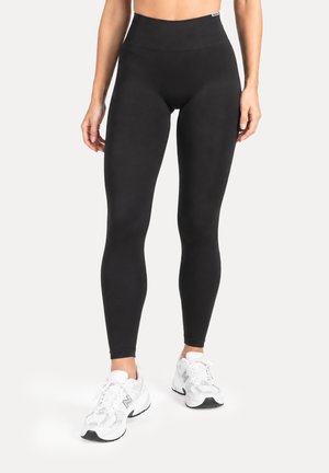 Mujer con leggings negros de talle alto y zapatillas deportivas blancas, de pie frente a un fondo liso y claro.