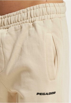 Beige joggingbukser med elastisk talje, sidelommer og en sort "PEGADOR"-logo. Fremstillet af blødt stof med en glat tekstur.