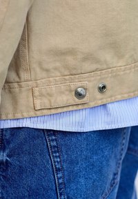 Beige jacket with snap button detail over blue striped shirt and blue denim jeans.