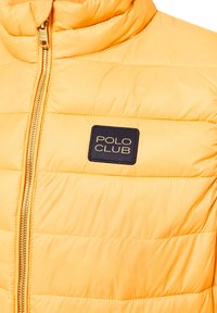Gul vadderad väst med dragkedja, med högt krage och en fyrkantig marinblå etikett med "POLO CLUB" i vit text. Slät textur.
