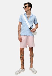 Camisa de manga corta azul claro texturizada, pantalones cortos rosas, sandalias deslizantes blancas y un bolso bandolera blanco. Tatuajes visibles en las piernas y los brazos.