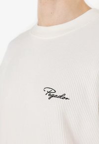 White ribbed sweater featuring a crew neck and black embroidered "Pegador" logo on the chest. Soft texture with a classic design.
