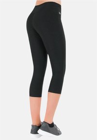 Schwarze Capri-Leggings aus elastischem Stoff, mit einem hohen Bund und einer glatten Textur. Tragen Sie sie mit grauen Sportschuhen.