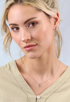 Silberne Halskette mit einem Tropfenanhänger, der einen kleinen Edelstein zeigt, getragen über einem beigen Shirt mit rundem Ausschnitt und Knopfdetails.