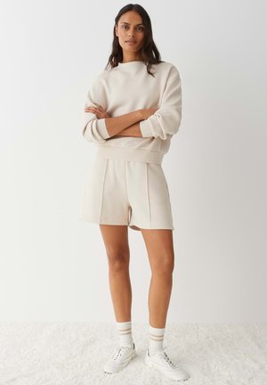 Frau steht mit verschränkten Armen, trägt ein cremefarbenes Sweatshirt, passende Shorts, weiße Turnschuhe und gestreifte Socken auf einem weißen Teppich.