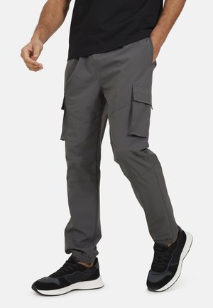 Mężczyzna w ciemnoszarych spodniach typu cargo z elastycznymi ściągaczami u dołu oraz czarnych sneakersach, stojący z jedną ręką w kieszeni, a drugą luźno opuszczoną.