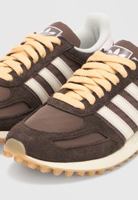 Ζεύγος καφέ αθλητικών παπουτσιών Adidas με μπεζ κορδόνια, λευκές τρεις ρίγες και υφή σόλας σε ανοιχτό φόντο.