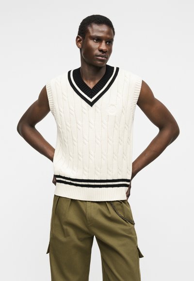 Άνδρας που φοράει λευκό αμάνικο πουλόβερ με πλέξη καλώδιο και μαύρες ρίγες, καθώς και χακί παντελόνι τύπου cargo, στέκεται με τα χέρια στη μέση.