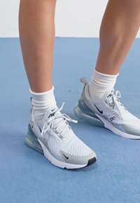 Fehér és szürke sportcipők, hálós textúrával, látható fűzőkkel és átlátszó légsólékkal. Fehér bokazoknival viselve.