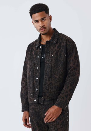 Uomo che indossa giacca e pantaloni con stampa leopardata marrone scuro e nera, camicia nera, in piedi davanti a uno sfondo chiaro e uniforme.