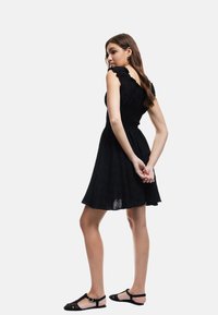 Schwarzes Kleid mit rüschierten Schulterträgern, strukturiertem Stoff, figurbetontem Oberteil und schwingendem Rock. Das Model trägt schwarze Ballerinas und steht seitlich.
