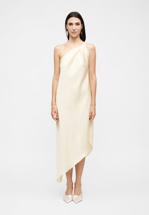 Vrouw met donker haar die een crèmekleurige asymmetrische jurk met één schouder draagt en witte puntige schoenen, staand tegen een effen lichte achtergrond.