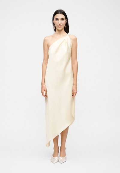 Femme aux cheveux foncés portant une robe asymétrique crème à une épaule et des chaussures blanches pointues, debout devant un fond clair uni.
