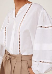 Valkoinen paita, jossa on nappilista ja pystysuorat leikkaukset. Pitkät, puhvimaiset hihat. Yhdistetty korkea vyötäröisiin beigeihin housuihin, joissa on solmittava vyö.