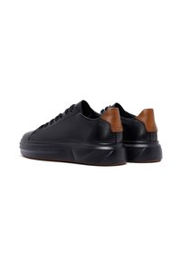 Un paio di sneaker in pelle nera con suole spesse e dettagli in pelle marrone sul tallone, viste dal retro su sfondo bianco.