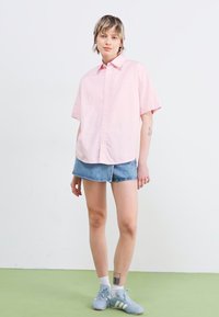 Camicia rosa a maniche corte con bottoni abbinata a pantaloni corti di jeans. La camicia ha una vestibilità rilassata; il modello indossa sneaker grigie e calze bianche.