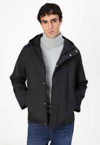 Μαύρο ελαφρύ μπουφάν με κουκούλα, κλείσιμο με κουμπιά και δύο μπροστινές τσέπες. Φορεμένο πάνω από ένα ανοιχτό γκρι ριμπ τζελ καθαρά.