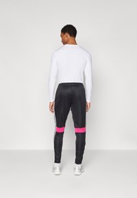 Schwarze Sporthose mit pinken Streifen, elastischem Bund und Seitenelementen; kombiniert mit einem langärmligen weißen Oberteil. Geschmeidige Textur.