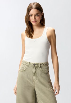 Νεαρή γυναίκα με κυματιστά καστανά μαλλιά, που φοράει λευκό τοπ και ψηλόμεσα μπεζ τζιν, στέκεται μπροστά σε απλό ανοιχτό φόντο.