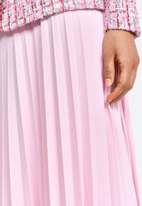 Jupe plissée en tissu rose doux, présentant une texture délicate. Une main effleurant doucement le tissu, mettant en valeur le tombé et le mouvement.