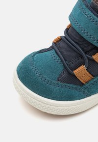 Baskets en daim teal avec des accents bleu marine, une sangle marron et des lacets élastiques. Présentent une semelle en caoutchouc blanc texturé et des détails cousus.