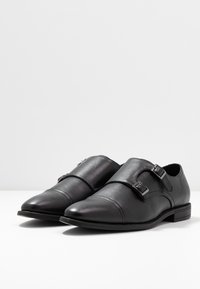 Chaussures en cuir noir avec un bout pointu, doubles brides à boucle et des détails en surpiqûre contrastés. Design à talon bas avec une texture lisse.