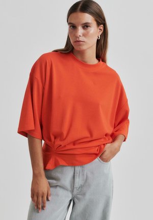 Tricou portocaliu, lejer, cu guler rotund și umeri căzuți, având un detaliu sub formă de nod la talie. Asortat cu pantaloni gri deschis.