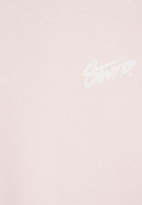Tissu rose clair avec un texte blanc pâle style coups de pinceau "Saint.", texture légèrement usée.