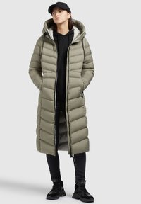 Padded long coat in olive green with a hood, zip closure, side pockets, and a textured surface. Worn over a black hoodie and jeans.