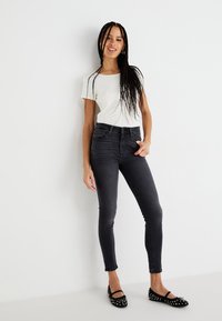 Skinny jeans in dark gray denim with a high waist, paired with a white fitted t-shirt and black studded flats, set against a white background.
