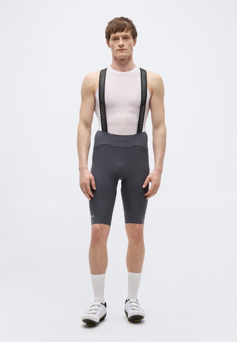 Jeune homme debout face à l'avant portant une couche de base cycliste blanche sans manches, un cuissard à bretelles noir, des chaussettes blanches et des chaussures de cyclisme blanches.