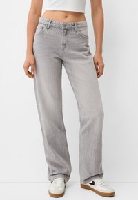 Jeans a gamba larga grigio chiaro realizzati in denim, con chiusura a bottone, tasche frontali e una texture sbiadita. Indossati con scarpe da tennis bianche.