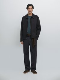 Zwarte zip-up jas over een teal shirt, donkere broek en bruine sneakers. Gladde textuur met een subtiel patroon op de stof.