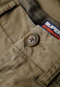 Khaki tkanina s jemnou texturou, s kulatým hnědým knoflíkem s vyraženým nápisem "Superdry" a částečně viditelnou námořnickou etiketou nad ním.