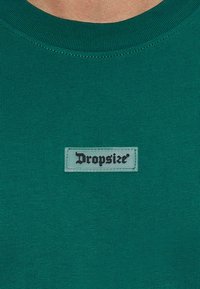 Camiseta de color verde oscuro con un escote acanalado y una etiqueta rectangular que presenta la palabra "Dropsize" en negro sobre un fondo claro. Textura de tela suave.