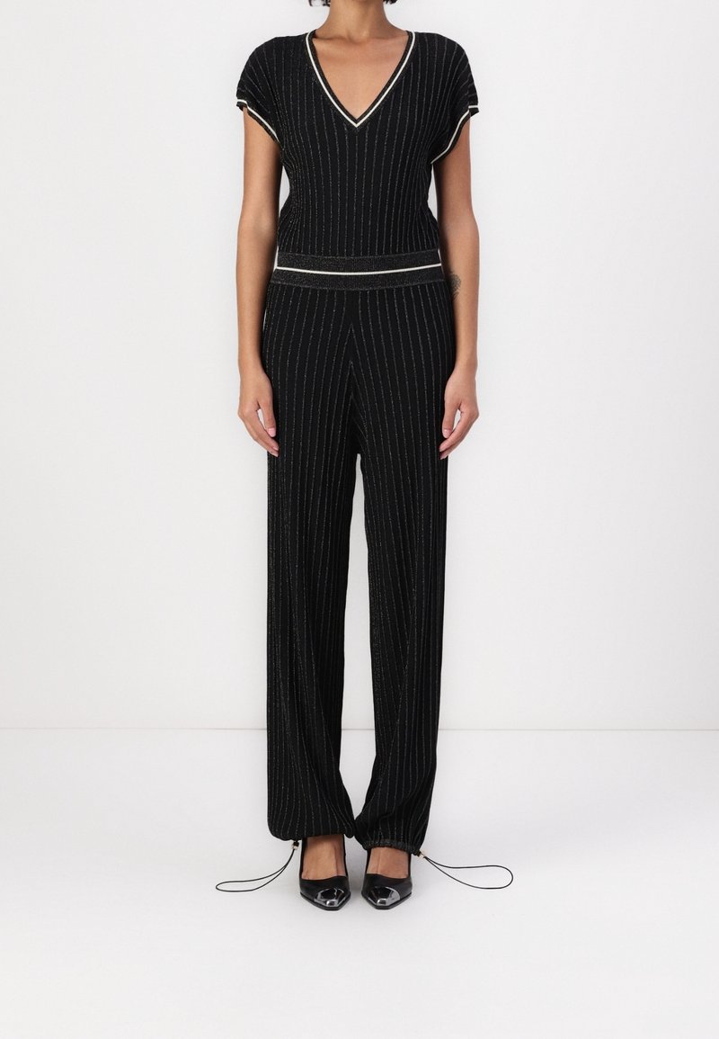 Femme portant une tenue en maille noire avec des rayures verticales argentées, un haut à col en V, un pantalon large avec des cordons aux chevilles, et des chaussures pointues noires.