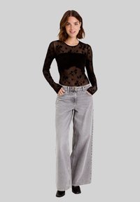 Haut noir transparent à manches longues avec un motif floral, associé à un jean large gris clair. Le modèle porte des bottines noires, les mains dans les poches.