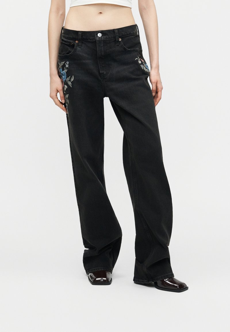 Άτομο που φοράει μαύρο τζιν παντελόνι ψηλόμεσο με φαρδιές παντελόνες και μπλε λουλουδένια κεντήματα στα πλαϊνά, μαζί με γυαλιστερά μαύρα παπούτσια με τετράγωνη μύτη, στέκεται σε λευκό πάτωμα.