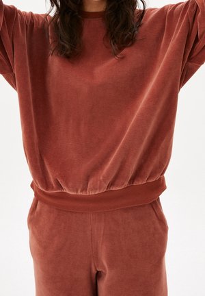 Personne portant un sweat-shirt ample en velours couleur rouille et un pantalon assorti, levant les bras avec la tête partiellement hors du cadre.